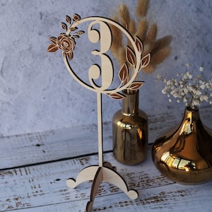 May include: Wooden table number with a floral design, featuring the number "3" in a circle. The table number is on a stand with three legs and is displayed on a white wooden surface with two gold vases.
