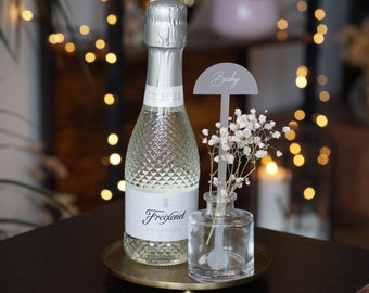 May include: A clear glass bottle of Freixenet Prosecco with a gold metal coaster. A small glass vase with white baby's breath flowers and a gray acrylic place card with the name "Becky" is in front of the bottle.