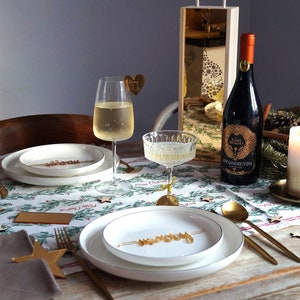 May include: A Christmas table setting with a white tablecloth with a green and red pattern. There are two place settings with white plates, gold cutlery, and brown napkins. A bottle of red wine with a gold label is on the table, along with a glass of white wine and a glass of champagne. There is a gold candle holder with a white candle in the center of the table.