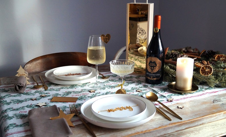 May include: A Christmas table setting with a white tablecloth with a green and red pattern. There are two place settings with white plates, gold cutlery, and brown napkins. A bottle of red wine with a gold label is on the table, along with a glass of white wine and a glass of champagne. There is a gold candle holder with a white candle in the center of the table.