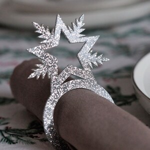 May include: Silver glitter star-shaped napkin ring. The ring is made of cardboard and has a snowflake design.
