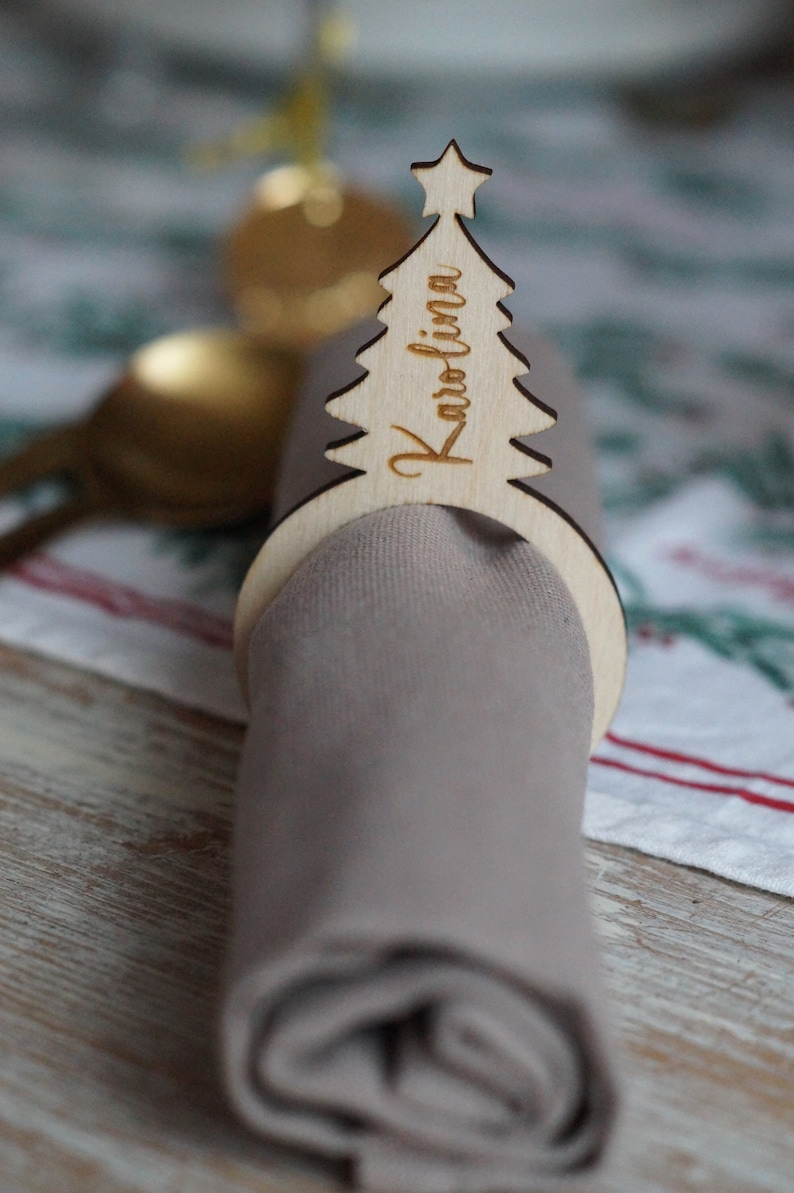 May include: A wooden Christmas tree napkin ring with the name "Karolina" engraved on it. The ring is holding a gray napkin.