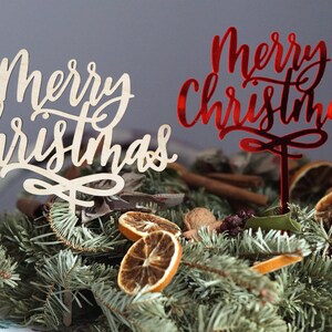 May include: Two wooden signs with the text "Merry Christmas" in a festive script font. One sign is natural wood and the other is painted red. The signs are placed in a decorative arrangement of pine branches, cinnamon sticks, and dried orange slices.