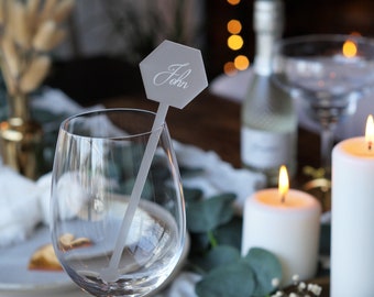 May include: A gray hexagonal place card with the name "John" written in white calligraphy, attached to a clear plastic stick, inserted into a clear wine glass. The glass is on a table with candles and greenery.