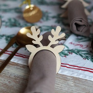 May include: A wooden reindeer napkin ring with a brown napkin. The napkin ring is a light brown color and has a simple design. The napkin is rolled up and placed inside the ring.