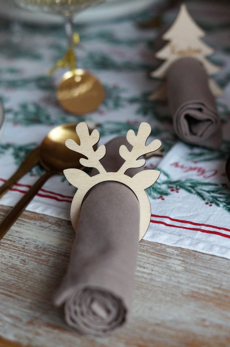 May include: A wooden reindeer napkin ring with a brown napkin. The napkin ring is a light brown color and has a simple design. The napkin is rolled up and placed inside the ring.