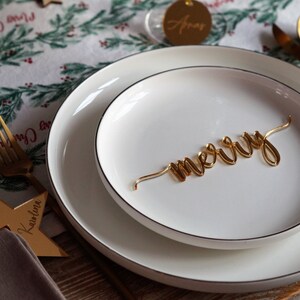 May include: A white dinner plate with a gold 'merry' sign on top of a smaller white plate. The plates are set on a table with a brown wooden surface. The table is covered with a white tablecloth with a green and red Christmas pattern.