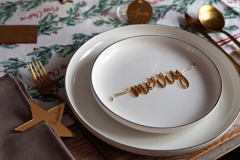 May include: A white dinner plate with a gold 'merry' sign on top of a smaller white plate. The plates are set on a table with a brown wooden surface. The table is covered with a white tablecloth with a green and red Christmas pattern.