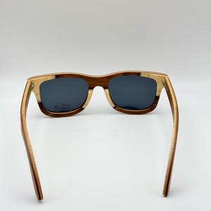 May include: Wooden sunglasses with a rectangular frame. The frame is a combination of light and dark brown wood, with dark lenses. The arms of the sunglasses are extended, showing the full length of the frame.
