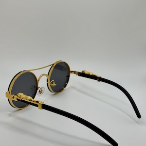 May include: Round sunglasses with gold frames and dark grey lenses. The frames have a double bridge and gold leopard detailing on the temples. The arms are black.