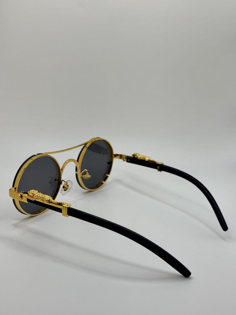 May include: Round sunglasses with gold frames and dark grey lenses. The frames have a double bridge and gold leopard detailing on the temples. The arms are black.