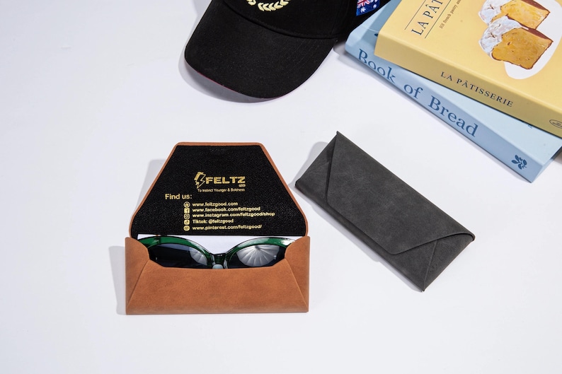 May include: A brown leather glasses case with green sunglasses inside, a black cap, and a grey glasses case. The open case displays the brand name "FELTZ" and social media details. Books are also visible in the background.