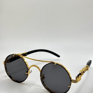 May include: Round sunglasses with a gold metal frame and dark grey lenses. The temples are black with gold decorative accents. The sunglasses have a double bridge and a sleek, modern design.