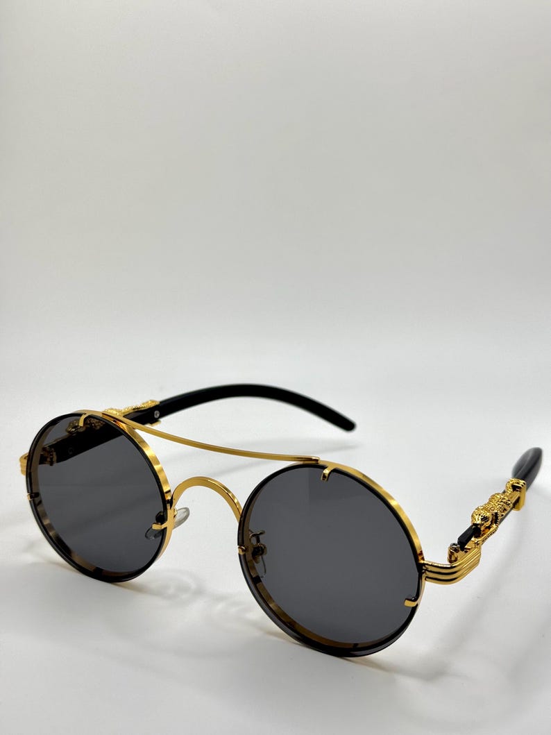 May include: Round sunglasses with a gold metal frame and dark grey lenses. The temples are black with gold decorative accents. The sunglasses have a double bridge and a sleek, modern design.