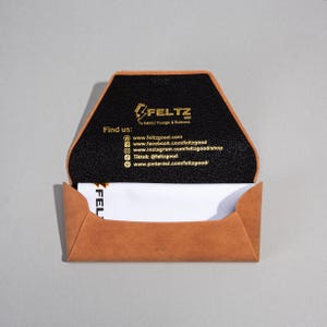 May include: Tan leather business card holder with a black interior, displaying the brand name "FELTZ GOOD" and social media details. A white business card is partially visible inside. The holder is open.