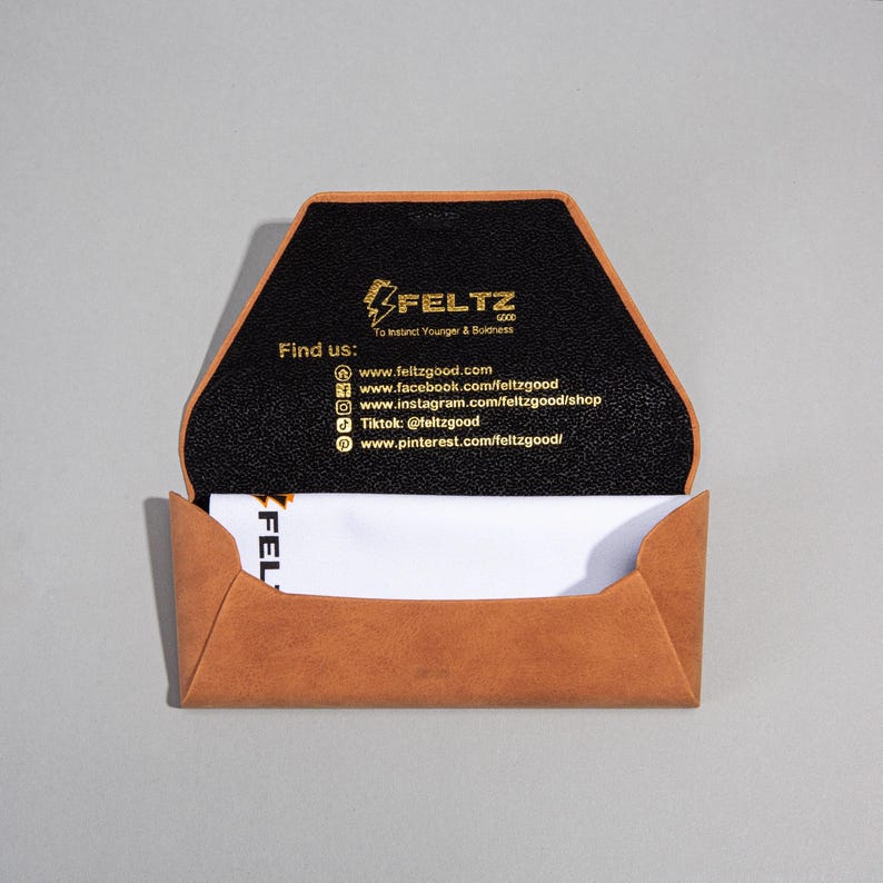 May include: Tan leather business card holder with a black interior, displaying the brand name "FELTZ GOOD" and social media details. A white business card is partially visible inside. The holder is open.