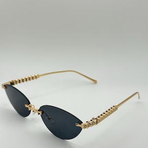 Puede incluir: Gafas de sol con montura dorada y lentes oscuras ovaladas. Las patillas presentan un diseño geométrico y las monturas tienen una estética elegante y moderna. Las gafas de sol están sobre un fondo blanco liso.