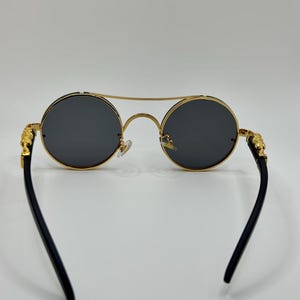 May include: Round sunglasses with gold frames and dark grey lenses. The temples are black and feature gold lion head accents. The bridge is a double bar design.