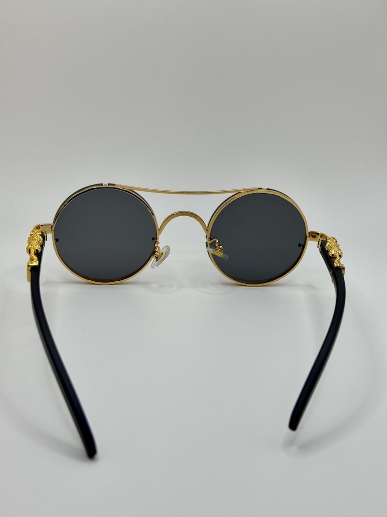 May include: Round sunglasses with gold frames and dark grey lenses. The temples are black and feature gold lion head accents. The bridge is a double bar design.