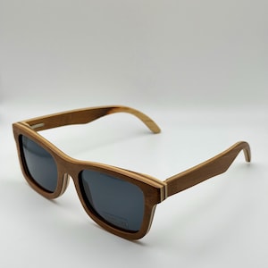 May include: Wooden sunglasses with dark lenses. The frames are a warm brown color with a layered design, and the arms curve gently. The sunglasses are set against a plain white background.