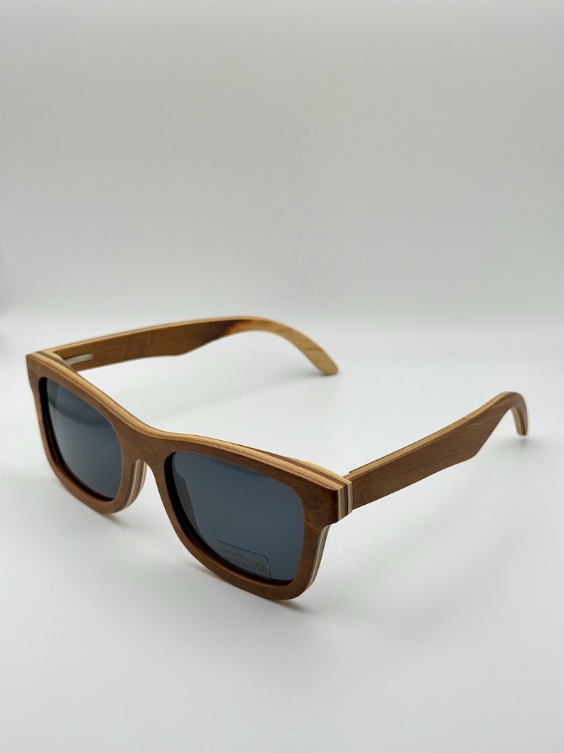 May include: Wooden sunglasses with dark lenses. The frames are a warm brown color with a layered design, and the arms curve gently. The sunglasses are set against a plain white background.