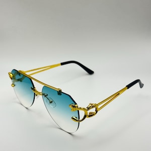 May include: Gold-framed sunglasses with a gradient blue lens. The sunglasses feature a double bridge and decorative gold accents on the temples. The arms are gold with black tips.