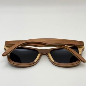 May include: Wooden sunglasses with a layered design. The frames are a combination of light and dark brown wood, with black lenses. The sunglasses are lying flat against a white background.