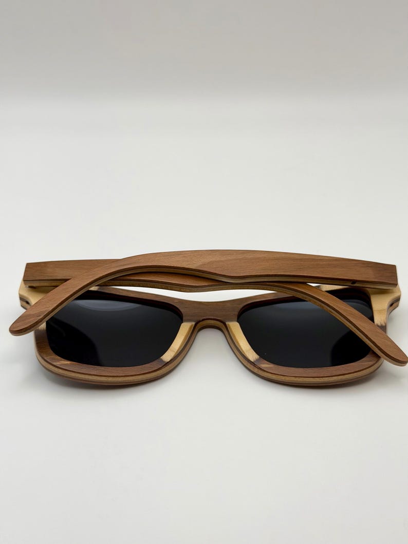 May include: Wooden sunglasses with a layered design. The frames are a combination of light and dark brown wood, with black lenses. The sunglasses are lying flat against a white background.