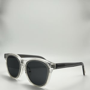 May include: Clear frame sunglasses with dark grey lenses. The sunglasses have a rounded shape and a thin, dark grey frame.