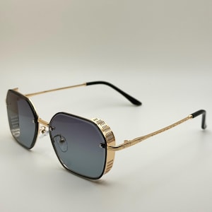 Puede incluir: Gafas de sol con montura dorada y un diseño geométrico único. Las lentes presentan un degradado azul y gris. Las patillas son finas y doradas, con terminales negros. Las monturas tienen un detalle texturizado distintivo en los laterales.