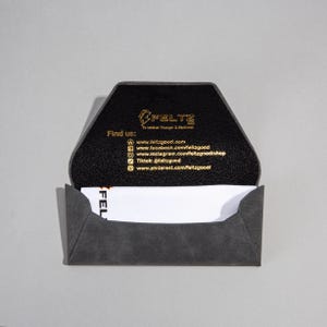 May include: A grey business card holder with a geometric design, open to reveal a white business card. The interior is black with gold text, including the brand name "FELTZ" and social media details.
