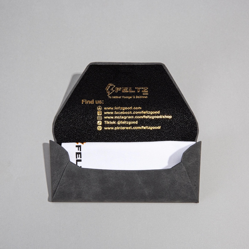 May include: A grey business card holder with a geometric design, open to reveal a white business card. The interior is black with gold text, including the brand name "FELTZ" and social media details.