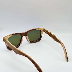 May include: Wooden sunglasses with a square frame and green lenses. The frame is a combination of light and dark wood tones. The arms are extended, showcasing the design and construction of the eyewear.