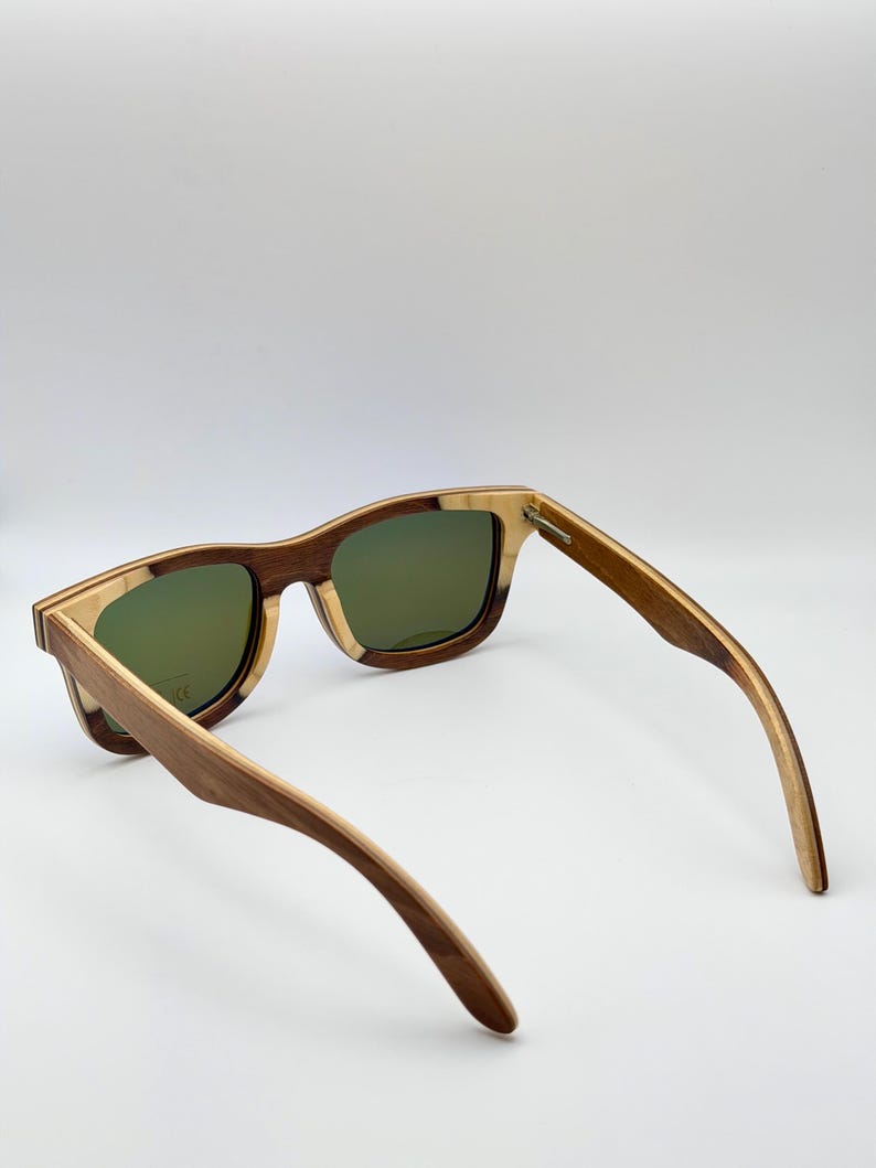 May include: Wooden sunglasses with a square frame and green lenses. The frame is a combination of light and dark wood tones. The arms are extended, showcasing the design and construction of the eyewear.