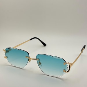 Könnte beinhalten: Randlose Sonnenbrille mit goldenem Rahmen und blau verlaufenden Gläsern. Die Gläser haben einen einzigartigen, gewellten Rand. Die Bügel sind goldfarben mit schwarzen Spitzen. Die Sonnenbrille ist vor einem weißen Hintergrund platziert.