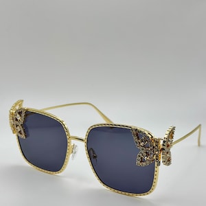 May include: Gold-tone square sunglasses with butterfly-shaped embellishments on the sides. The embellishments are covered in clear rhinestones. The sunglasses have dark gray lenses.