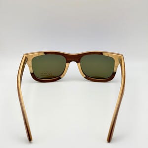 May include: Wooden-framed sunglasses with green lenses. The frames feature a combination of light and dark wood tones, creating a striped effect. The arms are a lighter wood color, and the lenses have a slight tint. The sunglasses are designed for UV 400 protection.