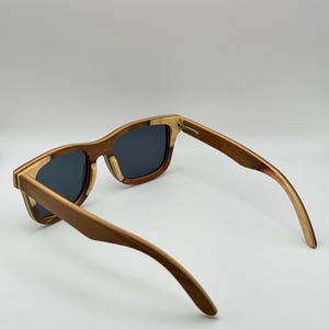 May include: Wooden sunglasses with a square frame and dark lenses. The frame is a mix of light and dark wood tones, with a natural grain pattern. The arms curve gently, and the sunglasses are set against a white background.