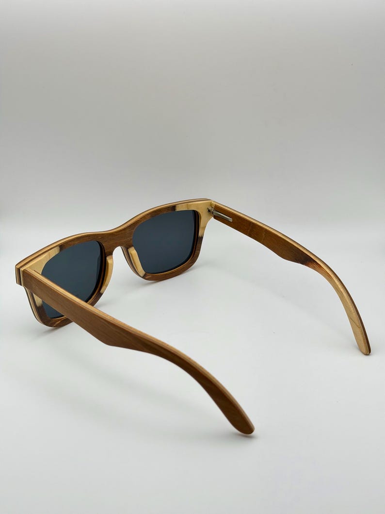 May include: Wooden sunglasses with a square frame and dark lenses. The frame is a mix of light and dark wood tones, with a natural grain pattern. The arms curve gently, and the sunglasses are set against a white background.