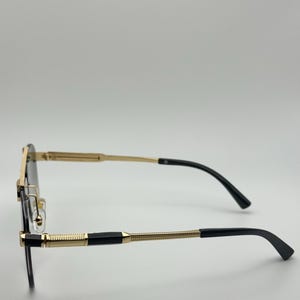 May include: Rimless spectacles with a gold frame and black arms. The glasses have a rectangular shape and a decorative gold pattern on the arms. The lenses are dark, and the overall design is sleek and modern.