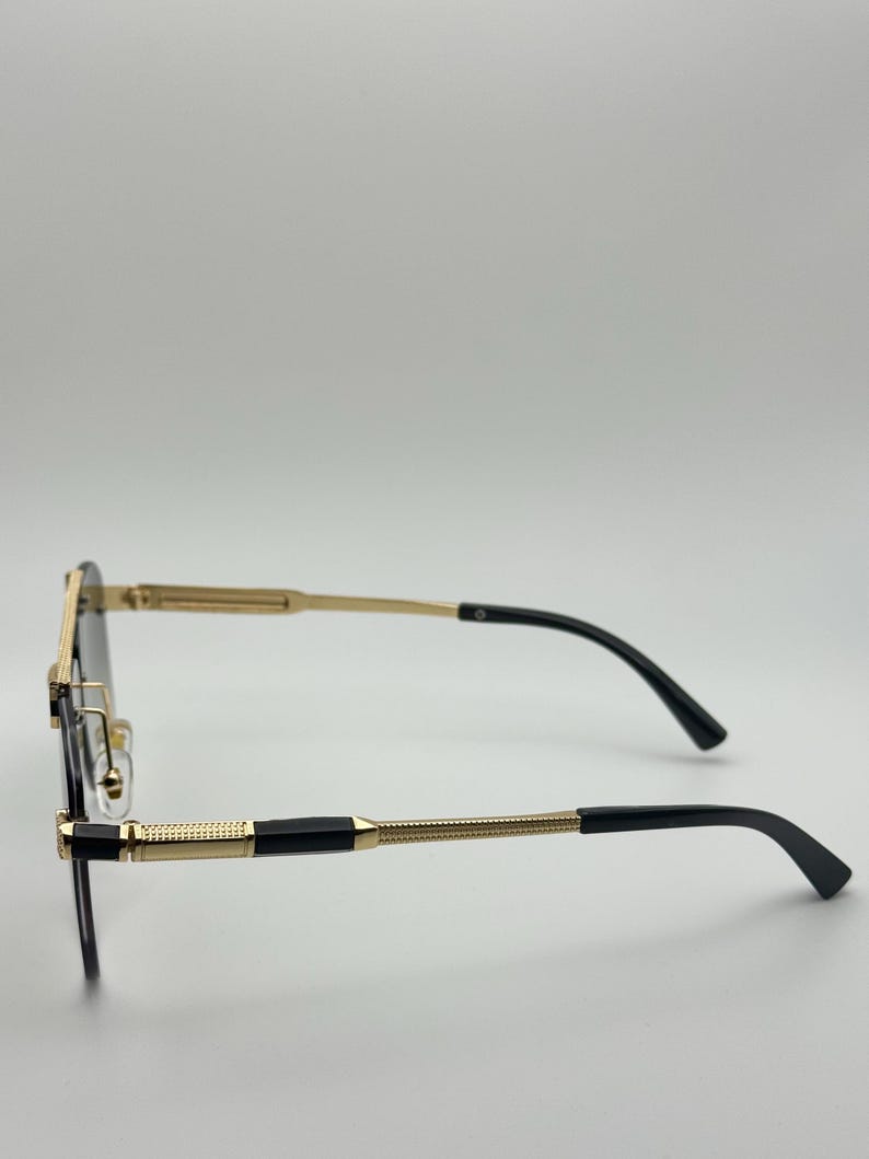 May include: Rimless spectacles with a gold frame and black arms. The glasses have a rectangular shape and a decorative gold pattern on the arms. The lenses are dark, and the overall design is sleek and modern.