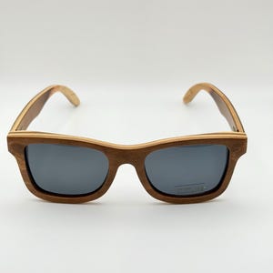 May include: Wooden-framed sunglasses with dark gray lenses. The sunglasses have a classic rectangular shape and a natural wood grain finish. The arms of the sunglasses are also made of wood.