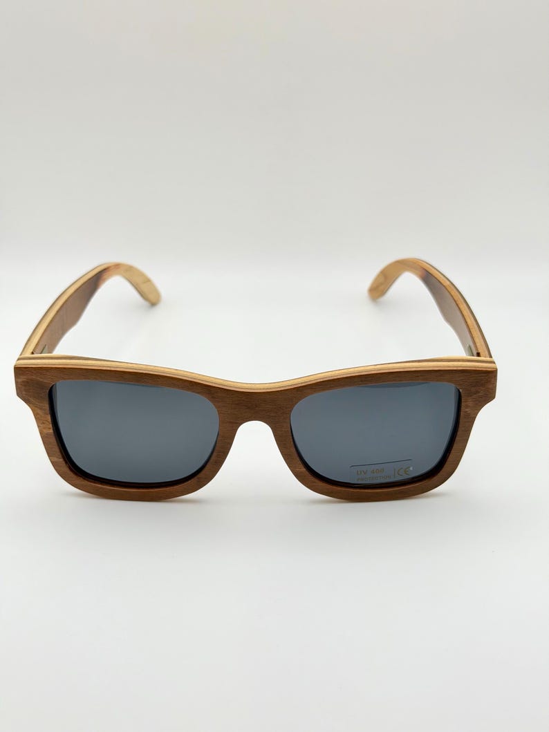 May include: Wooden-framed sunglasses with dark gray lenses. The sunglasses have a classic rectangular shape and a natural wood grain finish. The arms of the sunglasses are also made of wood.