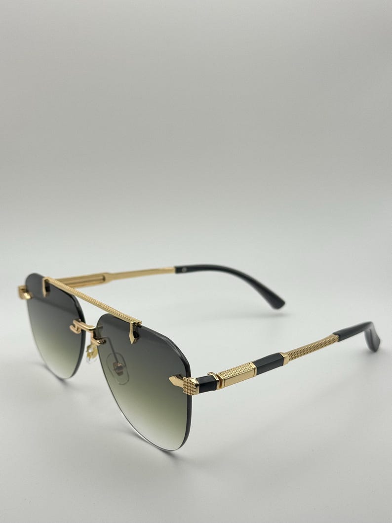 May include: Gold-rimmed sunglasses with a gradient lens. The sunglasses have a double bridge and black temples. The lenses transition from dark green to clear. The temples have a textured design.