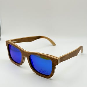 May include: Wooden-framed sunglasses with blue mirrored lenses. The sunglasses have a rectangular shape and a natural wood grain finish. The lenses reflect a bright blue color, and the frames are a light brown color.