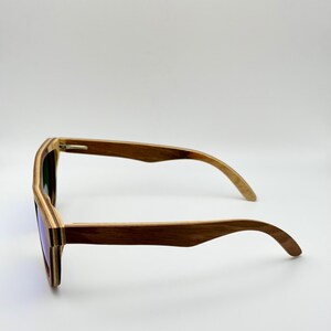 May include: Wooden sunglasses with a layered design. The frames are a mix of light and dark brown wood, and the lenses have a purple tint. The sunglasses are shown from a side profile.