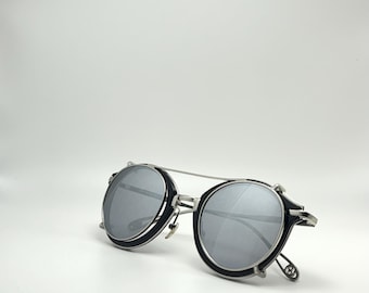Könnte beinhalten: Runde Sonnenbrille mit silbernem Rahmen und schwarzen Rändern. Die Gläser sind silberfarben reflektierend. Die Sonnenbrille hat einen Doppelsteg und ein einzigartiges Scharnierdesign.