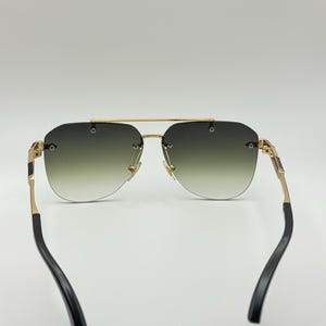 May include: Rimless sunglasses with a gold frame and gradient green lenses. The temples are gold with black accents. The sunglasses have a classic aviator shape.