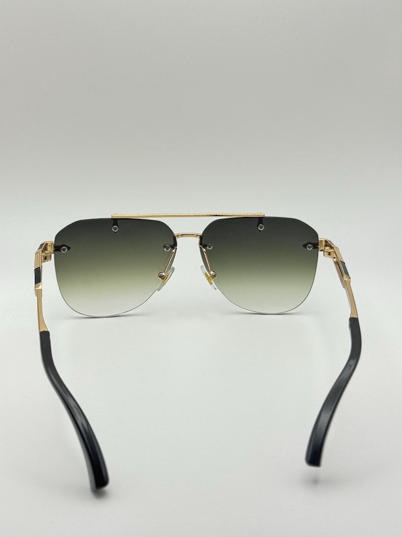 May include: Rimless sunglasses with a gold frame and gradient green lenses. The temples are gold with black accents. The sunglasses have a classic aviator shape.