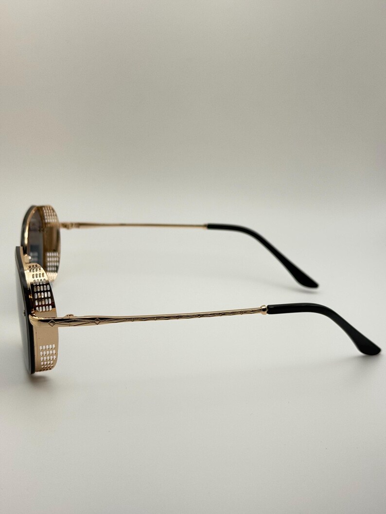 Pode incluir: &Oacute;culos de sol dourados com lentes e hastes pretas. As arma&ccedil;&otilde;es t&ecirc;m um padr&atilde;o decorativo em volta das lentes e nas hastes. As hastes s&atilde;o finas e t&ecirc;m pontas pretas. Os &oacute;culos de sol est&atilde;o em um fundo branco.